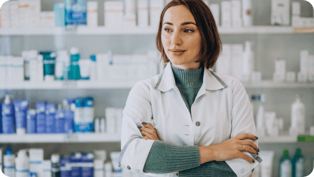 Uma farmacêutica de braços cruzados em frente à prateleira de remédios